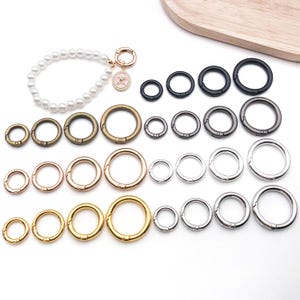 May include: An assortment of metal spring rings in various sizes and finishes, including gold, silver, bronze, and black. A pearl bracelet with a charm and a lobster clasp is also present. These rings are suitable for jewellery making and crafting.