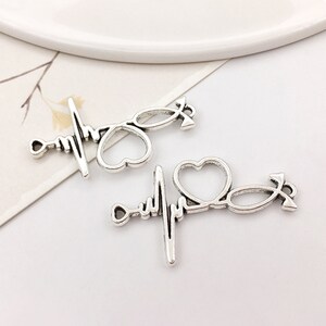 May include: Two silver-toned metal charms in the shape of a stethoscope, heart, and heartbeat line. The charms are perfect for jewelry making or crafting.