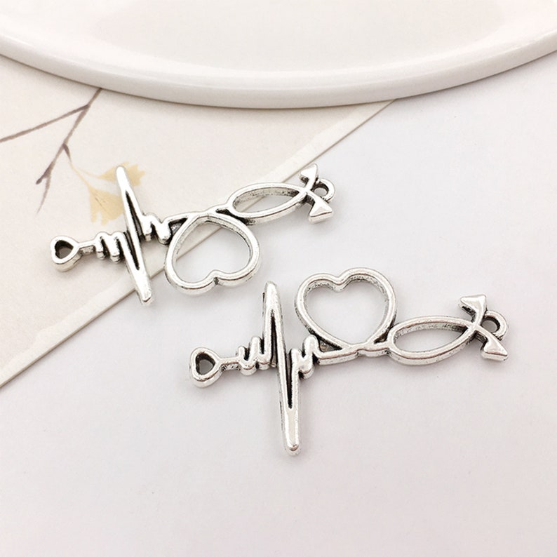 May include: Two silver-toned metal charms in the shape of a stethoscope, heart, and heartbeat line. The charms are perfect for jewelry making or crafting.