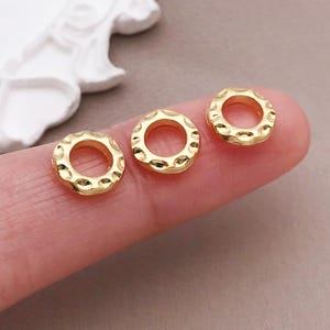 Pu&ograve; includere: Tre piccole perline ad anello dorate e strutturate. Ogni perlina ha una forma circolare con un foro al centro e un motivo ripetuto di piccole rientranze sul bordo esterno. Le perline sono esposte su una superficie rosa chiaro.