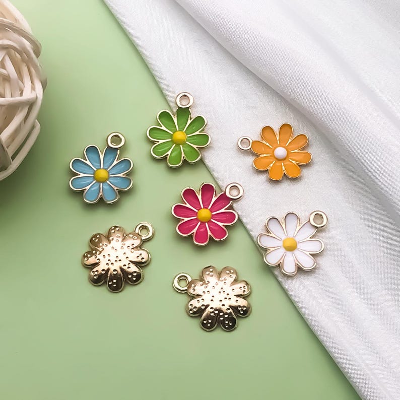 May include: Seven different flower charms with gold metal settings. The charms are in the shapes of daisies with different colored enamel centers and petals. The colors include green, blue, orange, pink, and white.