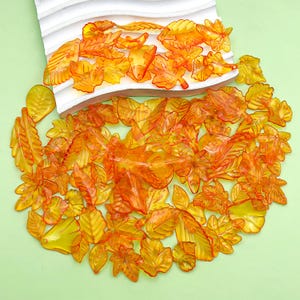 Könnte beinhalten: Eine Sammlung durchscheinender, orangefarbener und gelber blattförmiger Dekorationen. Die Blätter variieren in Größe und Form, wobei einige detaillierter erscheinen als andere. Sie sind auf einer hellgrünen Oberfläche angeordnet, wobei einige Blätter auf einer weißen, wellenförmigen Plattform liegen.