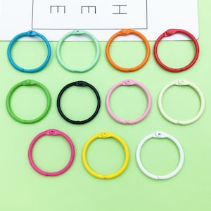 Puede incluir: Una colección de doce anillas de encuadernación de colores en varios tonos, incluyendo azul, verde, naranja y rojo. Las anillas están dispuestas sobre una superficie verde claro, con un fondo blanco en la parte superior. Cada anilla es de metal y tiene una abertura con bisagra.