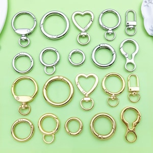 Puede incluir: Una variedad de llaveros en tonos plateados y dorados. Los llaveros incluyen diseños redondos, en forma de corazón y con cierre. El fondo es de color verde claro.
