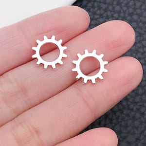 May include: A pair of silver-toned metal earrings shaped like gears or sunbursts. The earrings are small and delicate, with a simple design.