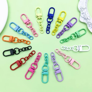 Könnte beinhalten: Eine Sammlung farbenfroher Metall-Drehhaken mit Kettengliedern. Die Verschlüsse sind in verschiedenen Farben erhältlich, darunter Rosa, Blau, Grün, Gelb, Orange und Schwarz. Die Ketten sind an den Verschlüssen befestigt und ebenfalls farblich passend.