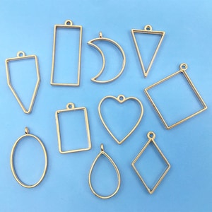 Könnte beinhalten: Zehn goldfarbene Metall-Anhänger-Rahmen in verschiedenen Formen, darunter ein Herz, ein Oval, ein Rechteck, ein Quadrat, eine Träne, ein Halbmond, ein Dreieck und zwei verschiedene Diamantformen. Die Rahmen sind alle offen und bereit, mit Harz oder anderen Materialien gefüllt zu werden.