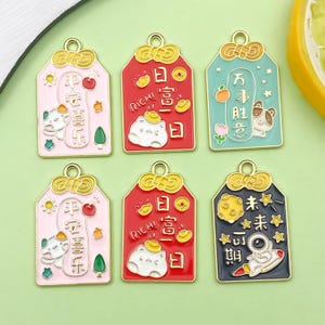 Puede incluir: Seis coloridos dijes de esmalte con detalles dorados. Los dijes presentan varios diseños, incluyendo un gato con sombrero dorado, un oso y un astronauta en un cohete. Cada dije tiene caracteres chinos y texto en inglés. Los dijes son en tonos rosa, rojo, azul y negro.