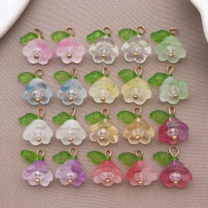 Peut inclure: Une collection de breloques en forme de fleurs colorées. Chaque breloque présente une fleur translucide de différentes couleurs, dont le rose, le bleu, le jaune et le vert, avec une bélière dorée et une feuille verte. Pour la création de bijoux.