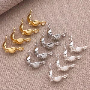 May include: A selection of gold and silver-toned metal jewellery findings, displayed on a textured, light brown surface. The findings have a curved, abstract shape with a small loop at the top, ideal for jewellery making.