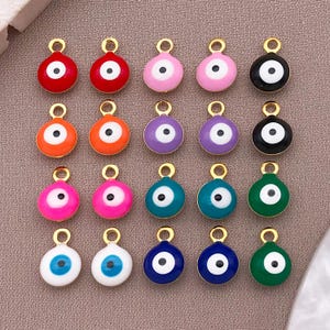May include: A collection of colorful enamel evil eye charms in various colors, including red, pink, orange, purple, blue, and green. Each charm has a gold-colored loop at the top for attaching to jewelry. The charms have a white iris and black pupil.