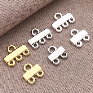 May include: Six metal jewellery clasps in gold and silver tones. Each clasp has a rectangular bar with three small loops on one side and a single loop on the other. The clasps are arranged on a textured, neutral-coloured surface.