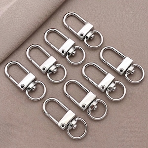May include: Eight silver keychains with a swivel clasp and a spring-loaded gate. The keychains are arranged in a row on a light brown surface.