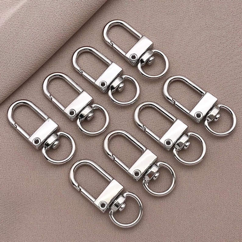 May include: Eight silver keychains with a swivel clasp and a spring-loaded gate. The keychains are arranged in a row on a light brown surface.