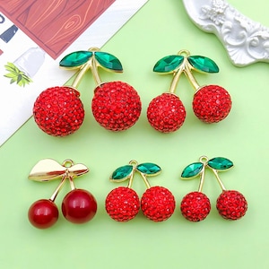 May include: Various cherry-shaped charms. Some have red, gem-covered cherries with gold stems and green gem leaves. Others have smooth, glossy red cherries. The charms are displayed on a light green surface.