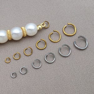 Puede incluir: Una variedad de anillas abiertas de color oro y plata en varios tamaños. Un hilo de perlas y cuentas doradas es visible en la esquina superior izquierda. Las anillas están dispuestas sobre una superficie de tono neutro.