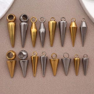 May include: An assortment of gold and silver-coloured metal pendants. The pendants are cone-shaped with a loop at the top for hanging. Some have a ring or ball at the top. The pendants are displayed on a neutral-toned surface.
