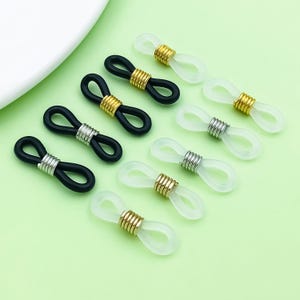 May include: A collection of eyeglass retainers in black and clear. The retainers have a loop design with metal and gold-colored accents. The background is a light green color.