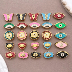 May include: A collection of colorful enamel charms in various shapes, including butterflies, eyes, hearts, and Hamsa hands. Each charm features gold-tone metal detailing and vibrant colors like pink, blue, red, and green. These charms are ideal for jewelry making.