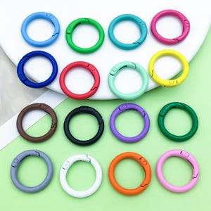 Puede incluir: Una colección de mosquetones redondos de colores en varios tonos, incluyendo azul, verde, rojo y rosa. Los mosquetones son de metal y tienen una puerta con resorte para una fácil fijación. La imagen muestra una variedad de colores.