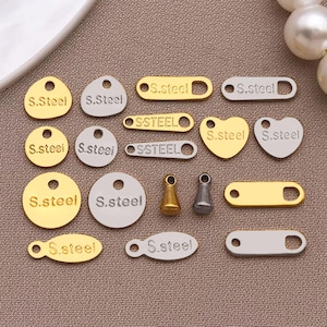 May include: A collection of gold and silver-toned metal tags and charms. The tags come in various shapes, including circles, ovals, hearts, and rectangles. Each tag is engraved with the text "S.Steel" or "SSTEEL".