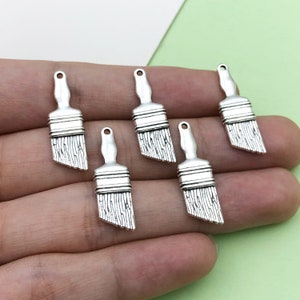 Puede incluir: Seis abalorios de pincel de plata, cada uno con una cabeza de pincel y un mango detallados. Estos abalorios son perfectos para añadir un toque de creatividad a las joyas o las manualidades.