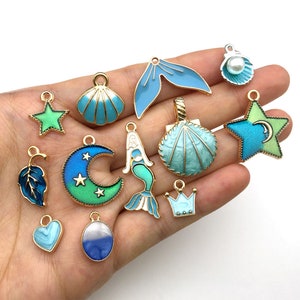 May include: Assortment of 13 gold-tone charms with blue and green enamel. Charms include a mermaid, seashells, stars, a crown, a leaf, a crescent moon, and a heart.