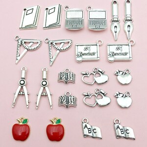 May include: A collection of silver charm pendants with school-themed designs, including books, apples, a protractor, a compass, a laptop, a pen, and a heart with an apple. The charms are perfect for adding a touch of school spirit to jewelry or crafts.