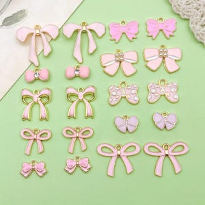 May include: A collection of pink and gold-toned bow charms in various designs. Some bows have pearl or rhinestone accents. The charms are on a light green surface, suitable for jewelry making or craft projects.