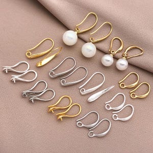 Op de afbeelding: Een assortiment gouden en zilverkleurige oorbelcomponenten, waaronder haken en oorbellen met pareldruppels. De haken variëren in stijl en maat, sommige met een eenvoudig gebogen ontwerp en andere met een decoratieve lus. De parels zijn wit en rond.