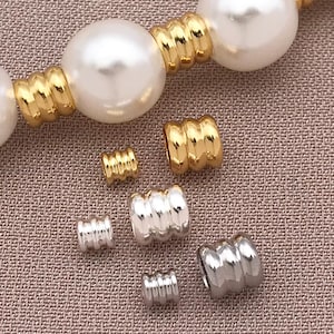 May include: Close-up of a pearl necklace with gold-colored ribbed spacers. Several gold and silver ribbed spacers are scattered around the necklace. The pearls are large and white.