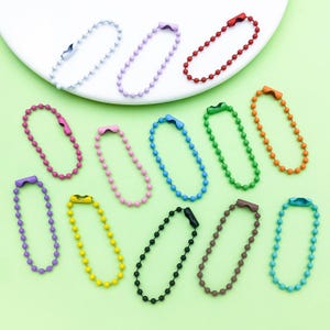 Puede incluir: Una colección de llaveros de cadena de bolas de colores, que incluyen blanco, morado, rojo, rosa, azul, verde, naranja, amarillo, negro, marrón y turquesa. Los llaveros están dispuestos sobre una superficie verde claro.