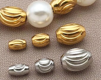 May include: Close-up of a necklace featuring large, white, round pearls and gold-colored, ridged beads. Several smaller gold and silver ridged beads are scattered below. The beads have a metallic sheen and are arranged on a neutral background.