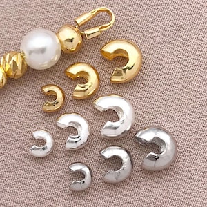 Könnte beinhalten: Eine Auswahl an gold- und silberfarbenen Schmuckzubehörteilen, darunter C-förmige Quetschhülsen und ein goldfarbener Verschluss. Ein Strang aus Perlen und goldenen Perlen ist ebenfalls zu sehen. Die Zubehörteile sind auf einer neutralfarbenen Stoffoberfläche angeordnet.