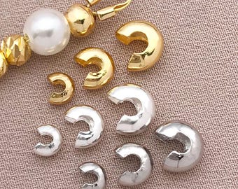 May include: An assortment of gold and silver-colored jewelry findings, including C-shaped crimp end findings and a gold-toned clasp. A pearl and gold bead strand is also visible. The findings are arranged on a neutral-toned fabric surface.