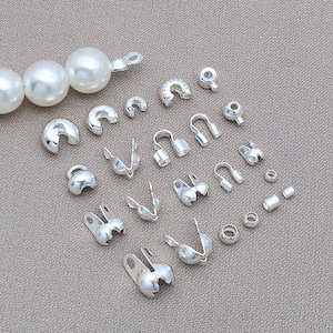 May include: An assortment of silver-colored jewelry findings, including clasps, crimp beads, and end caps. The findings are displayed on a neutral gray surface, with a strand of large white pearls visible at the top left.