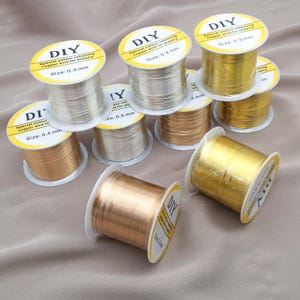 Op de afbeelding: Een verzameling spoelen DIY koperdraad voor het maken van sieraden. De spoelen zijn in verschillende metallic kleuren, waaronder goud, zilver en koper. Elke spoel heeft een label met de tekst "DIY" en maat informatie, variërend van 0,3 mm tot 0,5 mm.