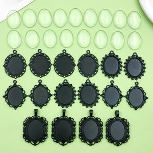 May include: A collection of black, oval pendant trays with decorative filigree borders, alongside clear, oval glass cabochons. The components are displayed on a pale green surface, suitable for jewellery making or crafts.