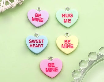 May include: Five heart-shaped charms with different Valentine's Day messages. The charms are pink, light blue, yellow, and green. The messages are "Be Mine", "Sweetheart", "Be Mine", "Hug Me", and "Be Mine".