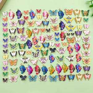 Puede incluir: Una colección de coloridos dijes de mariposa en varios tamaños y diseños. Los dijes presentan una gama de colores, incluyendo rosa, azul, amarillo y verde, con detalles metálicos dorados. Estos dijes decorativos son ideales para la fabricación de joyas y manualidades.