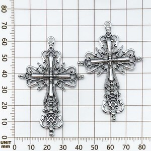 May include: Two ornate silver-colored crosses with intricate filigree detailing. Each cross features a loop at the top for hanging and measures approximately 3 inches in length. The design includes decorative flourishes and a central crossbar.