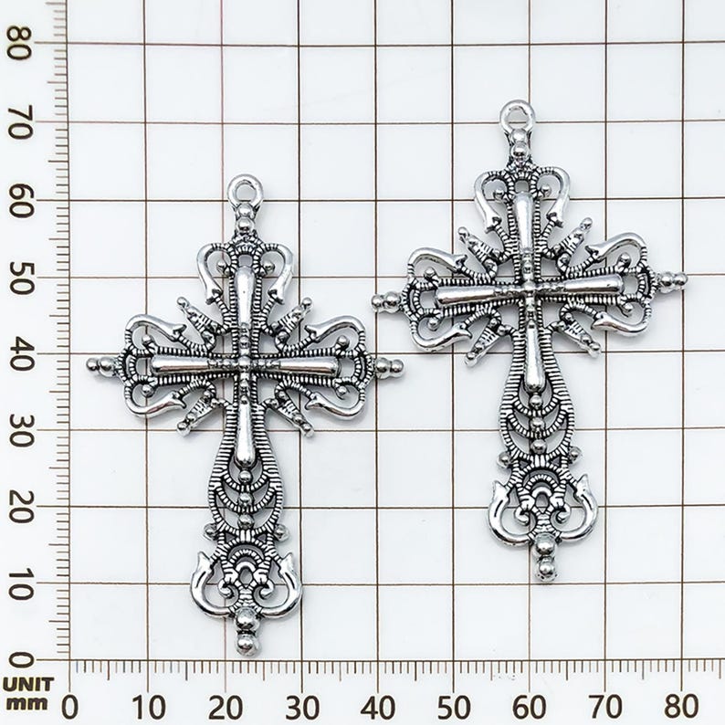 May include: Two ornate silver-colored crosses with intricate filigree detailing. Each cross features a loop at the top for hanging and measures approximately 3 inches in length. The design includes decorative flourishes and a central crossbar.