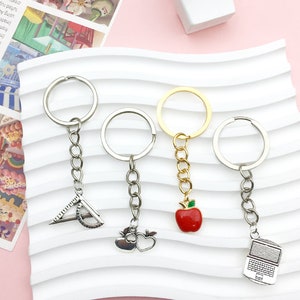 May include: Four silver keychains with different charms. The charms are a protractor, an apple, a heart, and a laptop. The keychains are on a white background.