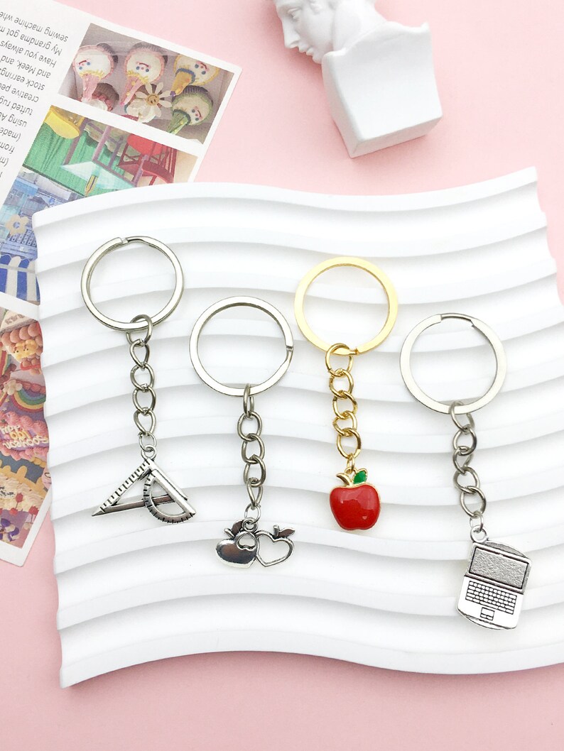 May include: Four silver keychains with different charms. The charms are a protractor, an apple, a heart, and a laptop. The keychains are on a white background.