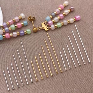 May include: A selection of jewellery-making components is presented. Beaded earrings with pastel beads and gold details are visible. Various silver and gold head pins of different lengths are arranged on a neutral-toned surface, ideal for crafting.