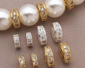 May include: An assortment of gold and silver-toned hoop-style jewelry with clear, sparkling stones. The jewelry is displayed near large, white, round beads, suggesting a necklace or bracelet design. The pieces vary in size and design.