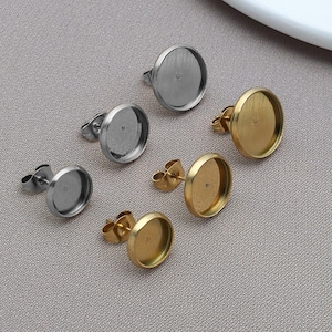 May include: A selection of earring posts in silver and gold colours. The posts have a flat, round base designed to hold a decorative element. The earrings are displayed on a neutral, light grey surface.