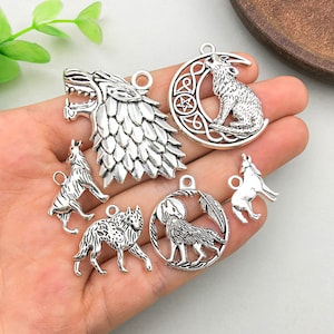 Puede incluir: Un conjunto de siete dijes de metal plateado con diseños de lobos. Los dijes incluyen una cabeza de lobo, un lobo aullando a la luna, un lobo sentado en una luna creciente y tres siluetas de lobos diferentes.