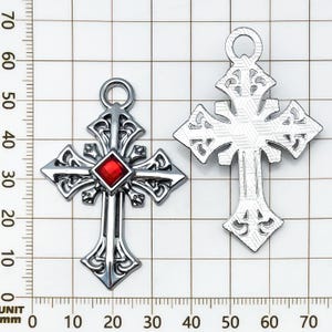 May include: Two silver-colored cross pendants. One cross features a red gemstone at the center. Both crosses have ornate, openwork designs and a loop at the top for hanging. The crosses are displayed against a grid background.