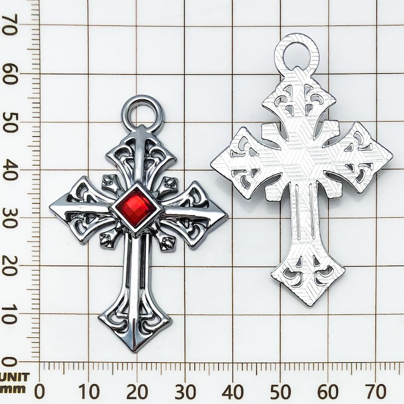 May include: Two silver-colored cross pendants. One cross features a red gemstone at the center. Both crosses have ornate, openwork designs and a loop at the top for hanging. The crosses are displayed against a grid background.